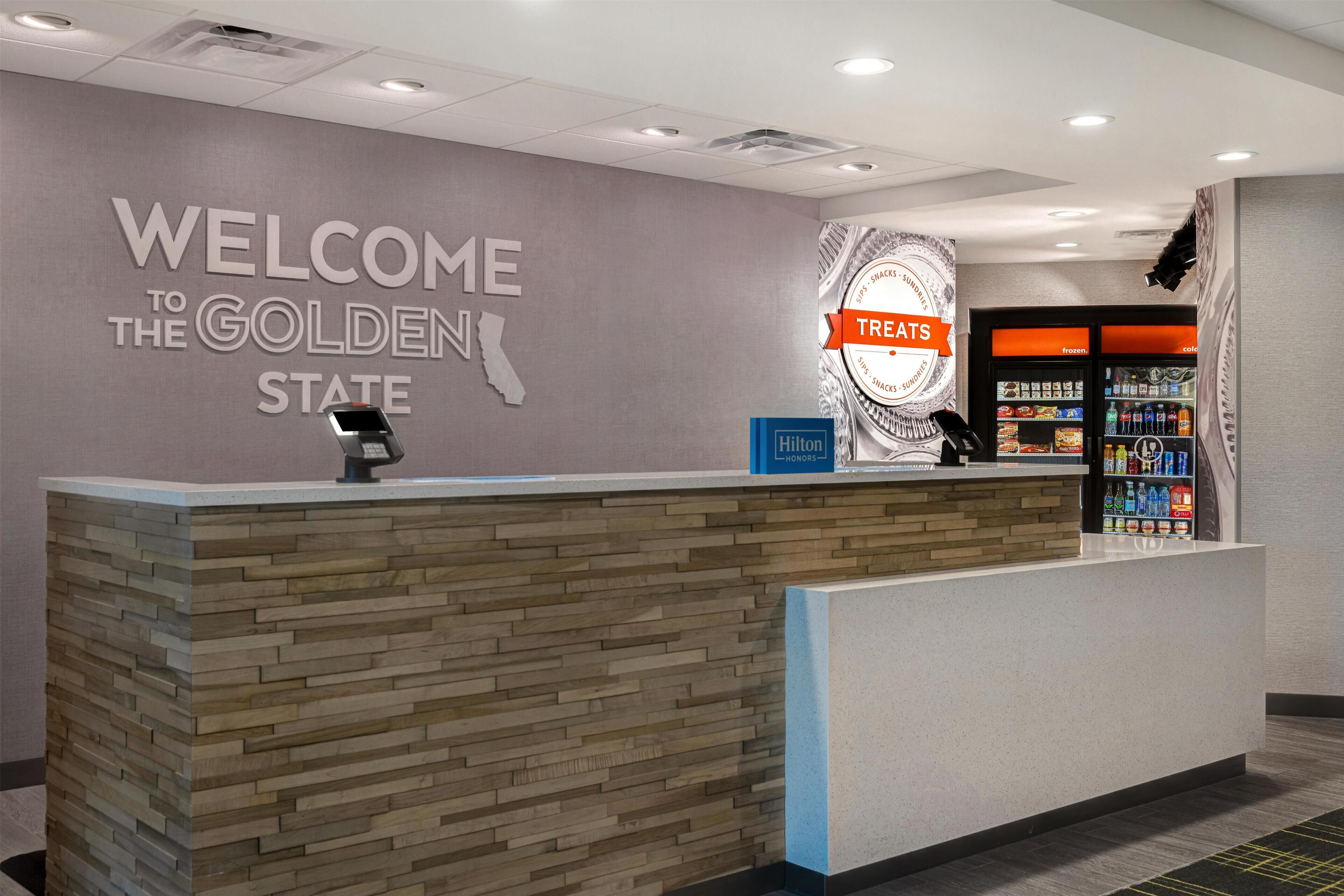 A reception desk with a 'Welcome to the Golden State' sign, a 'Treats' sign, and a minibar area.