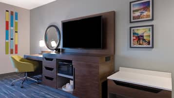 A hotel room with a flat-screen TV, a desk, a chair, a microwave, and a small shelf with cups.