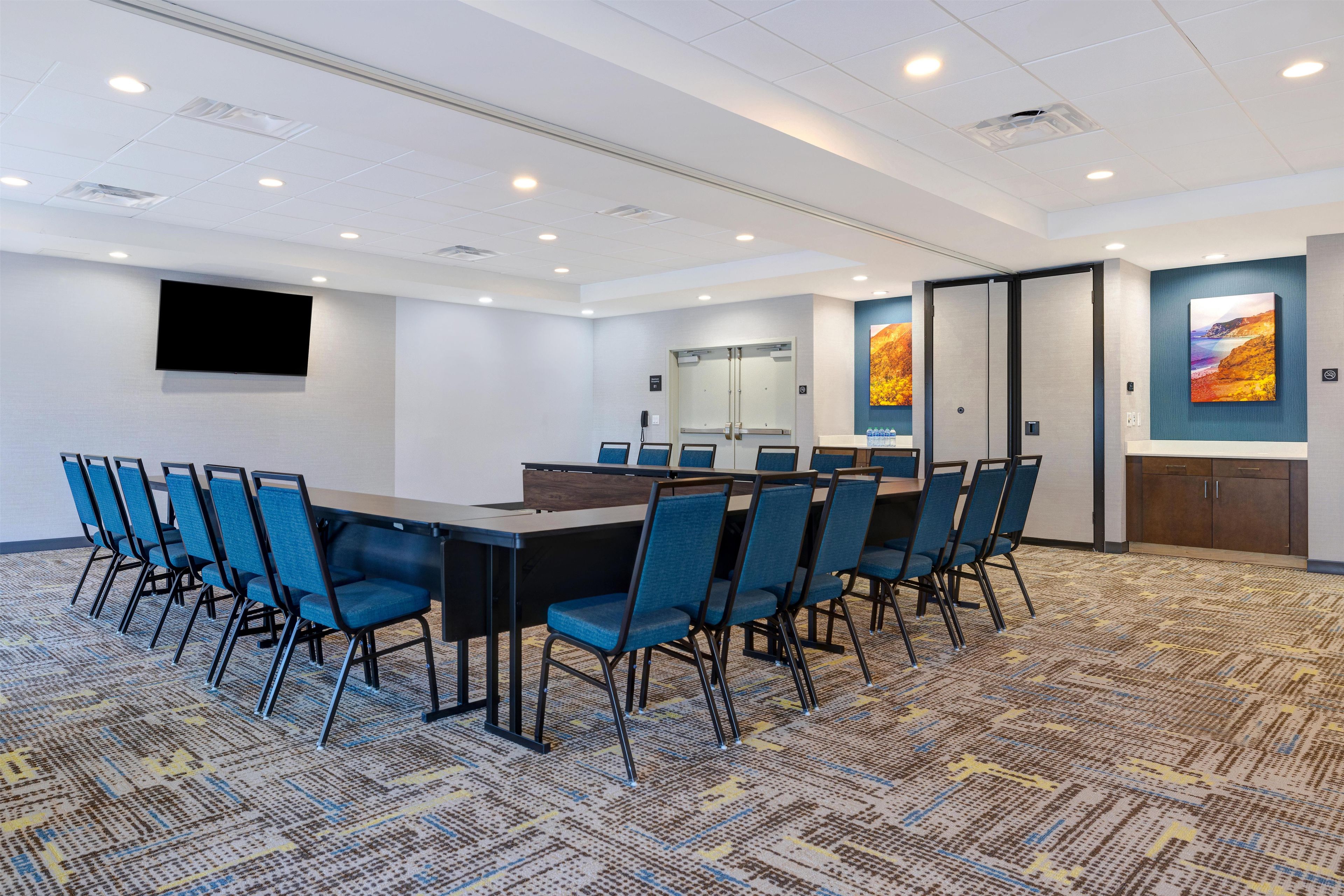 A conference room with a long table, blue chairs, a flat-screen TV, and a large painting on the wall.