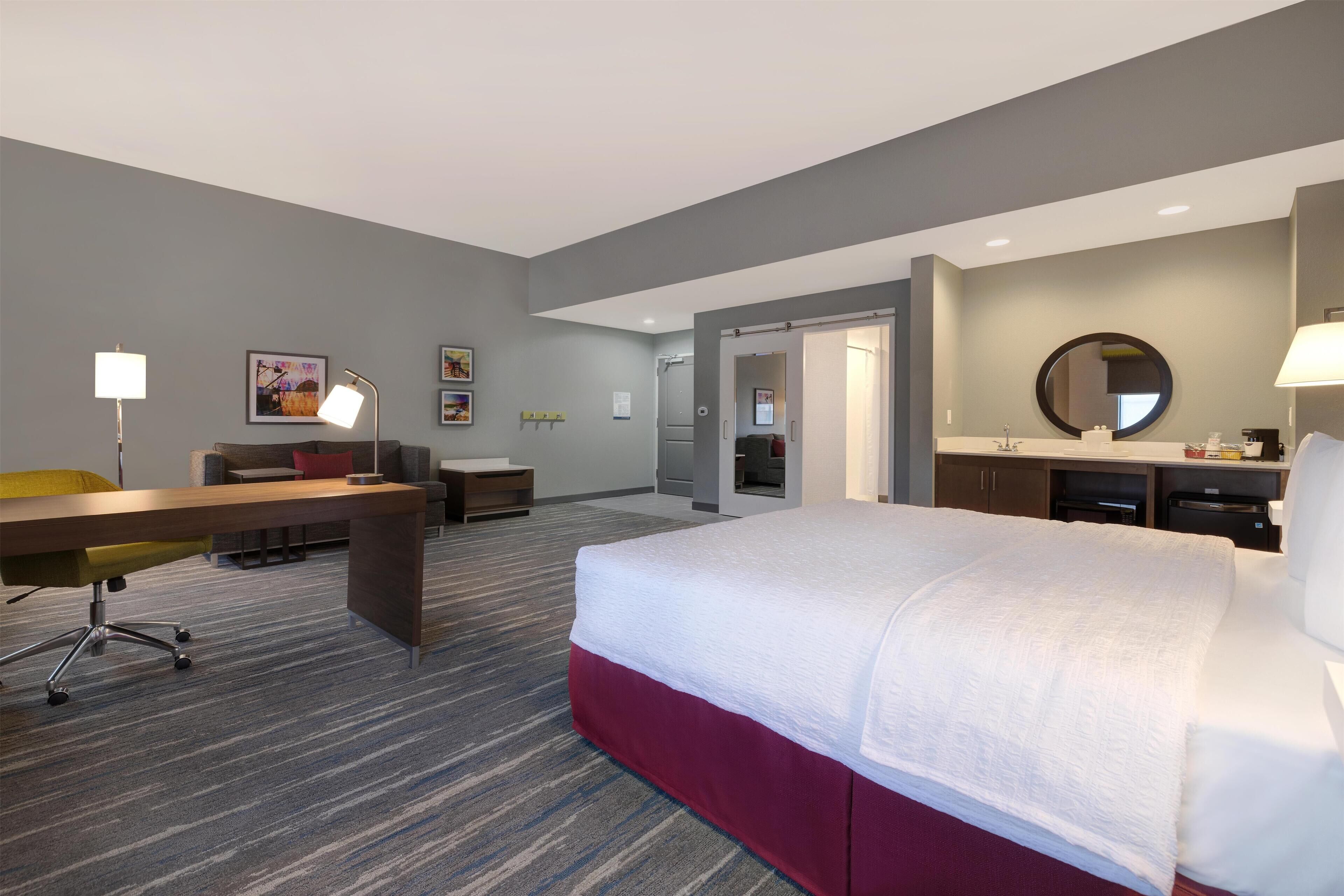 A hotel room with a bed, desk, chair, and a bathroom with a mirror.