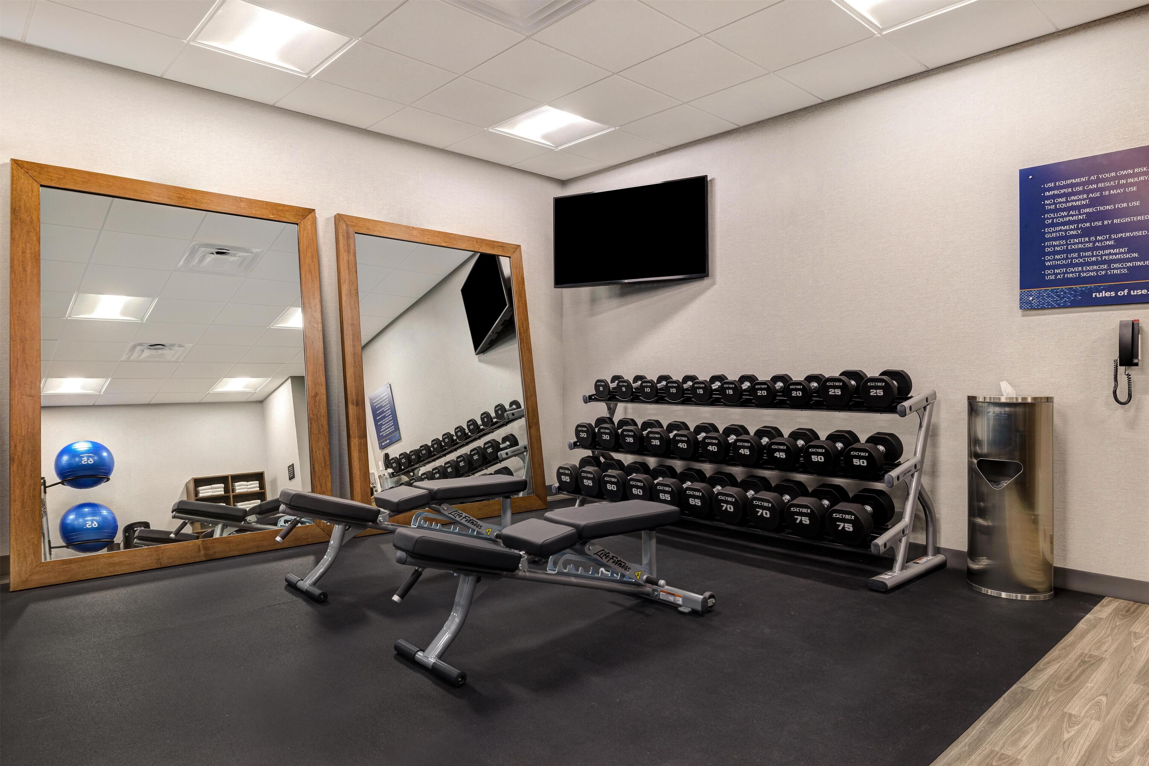 Une salle de sport équipée de matériel de musculation, de miroirs, d’un téléviseur et d’un panneau fixé au mur.