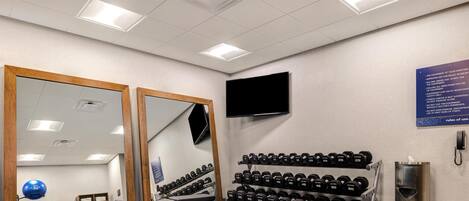 Une salle de sport équipée de matériel de musculation, de miroirs, d’un téléviseur et d’un panneau fixé au mur.
