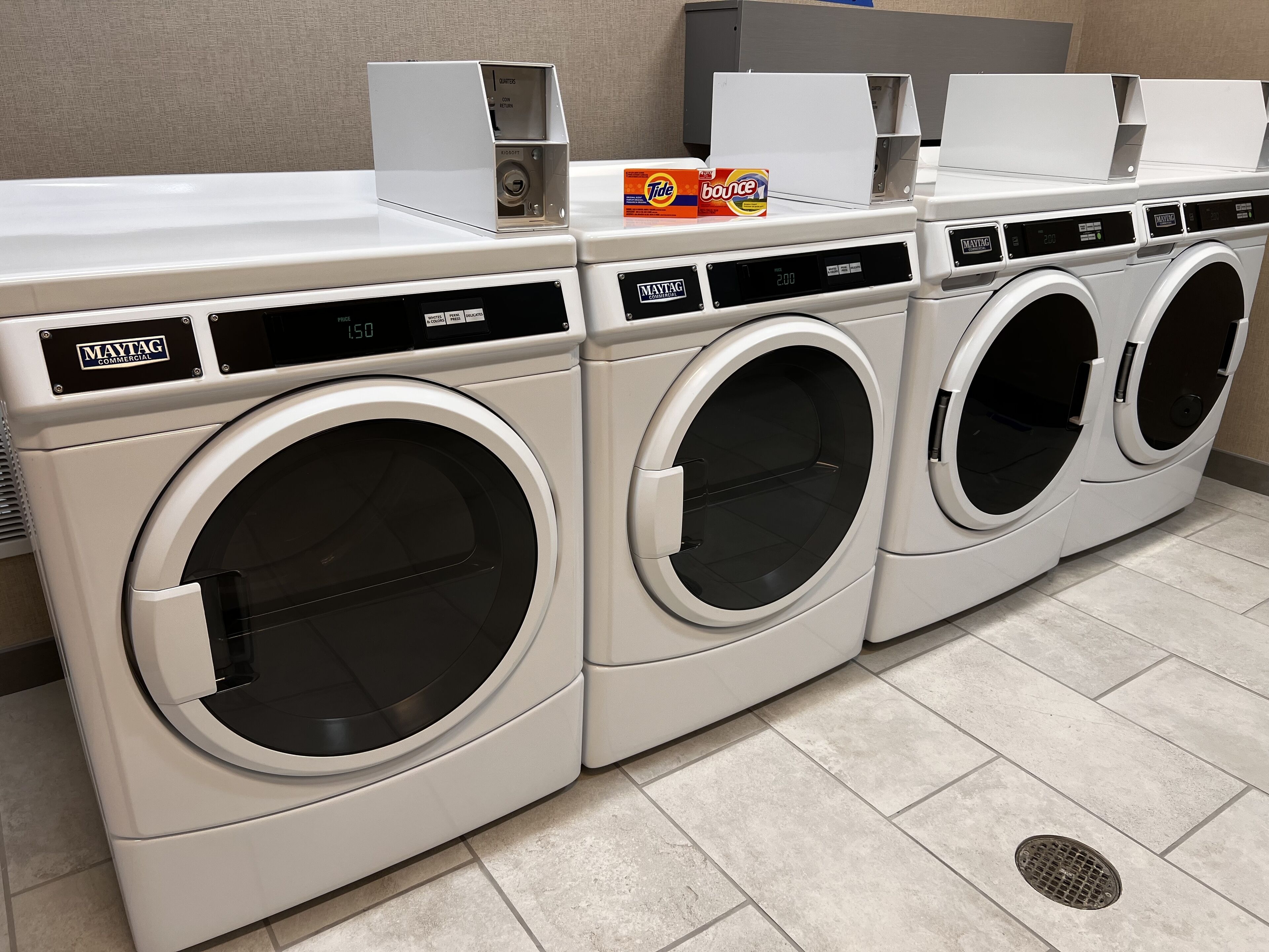 A row of white commercial washers with digital displays, a control panel, and a detergent box on top.