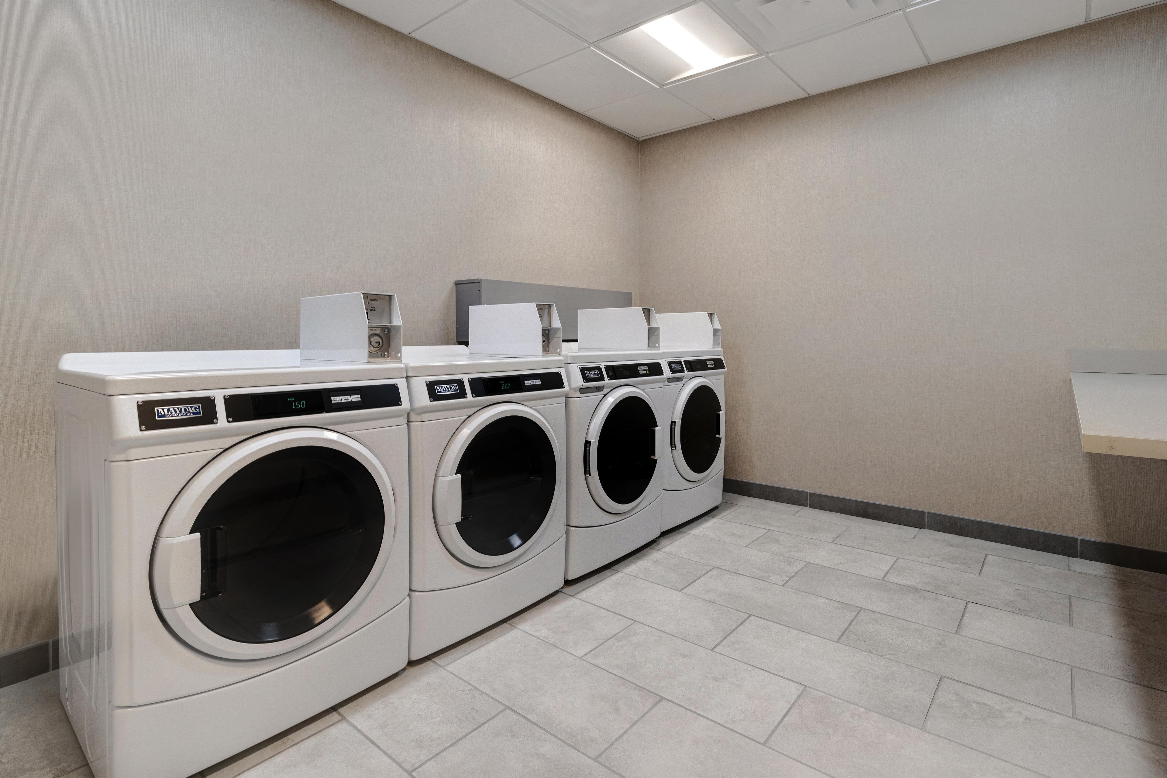 A laundry room with three front-loading washing machines and a dryer, a white countertop, and a tiled floor.