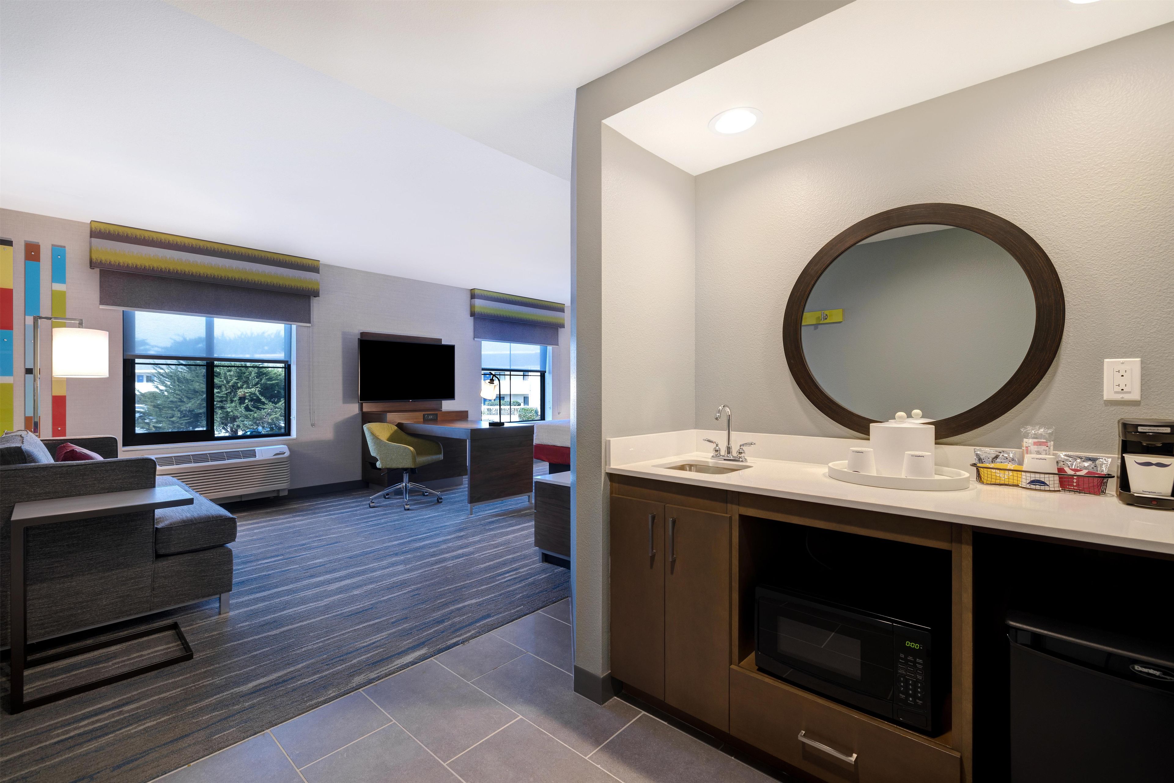 A hotel room with a sofa, a desk, a flat-screen TV, a large mirror, a sink, and a microwave.