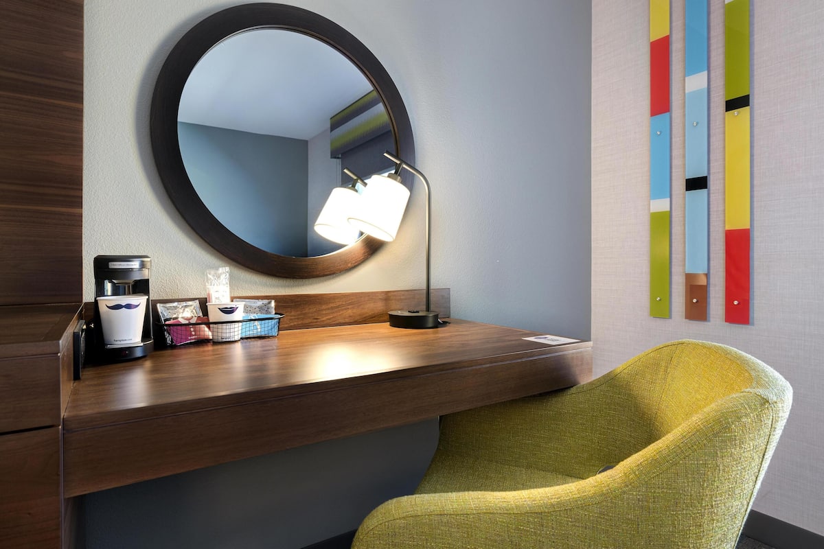 Een moderne badkamer met een ronde spiegel, een bureau met een lamp, een koffiezetapparaat en een stoel.