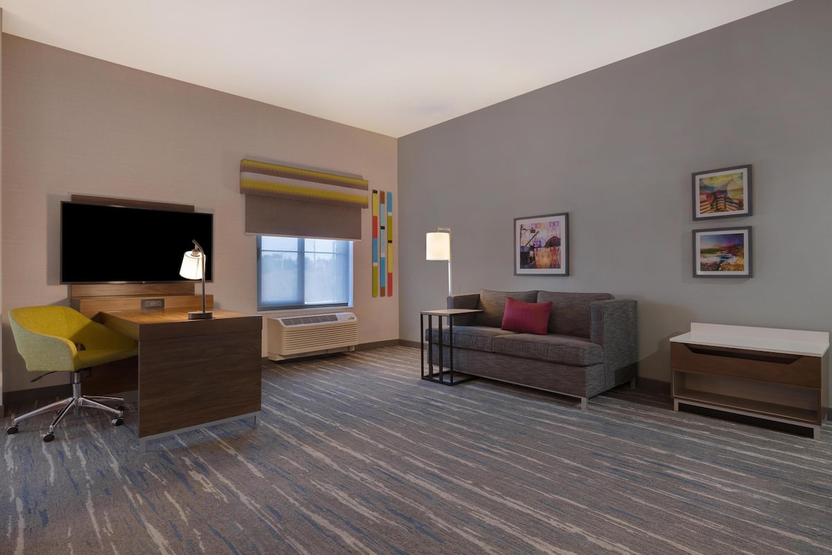 Een hotelkamer met een bureau, stoel, bank, televisie, airconditioning en kunstwerken aan de muren.