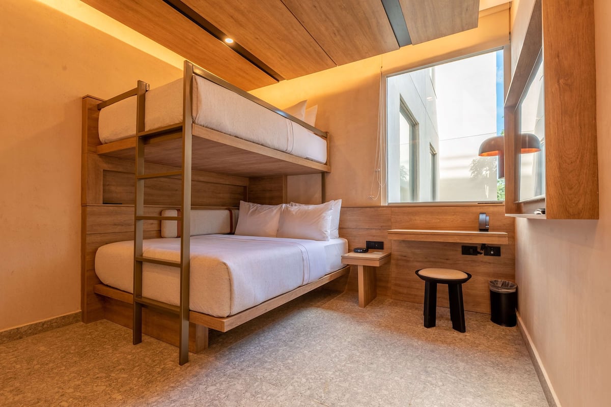 A bunk bed with a wooden ladder, a small stool, and a window with a view of the outside.