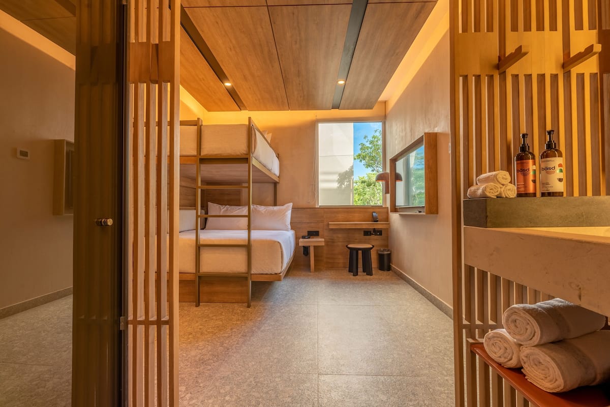 A compact hotel room with a bunk bed, a small desk, a window with a view, and a shelf with toiletries.
