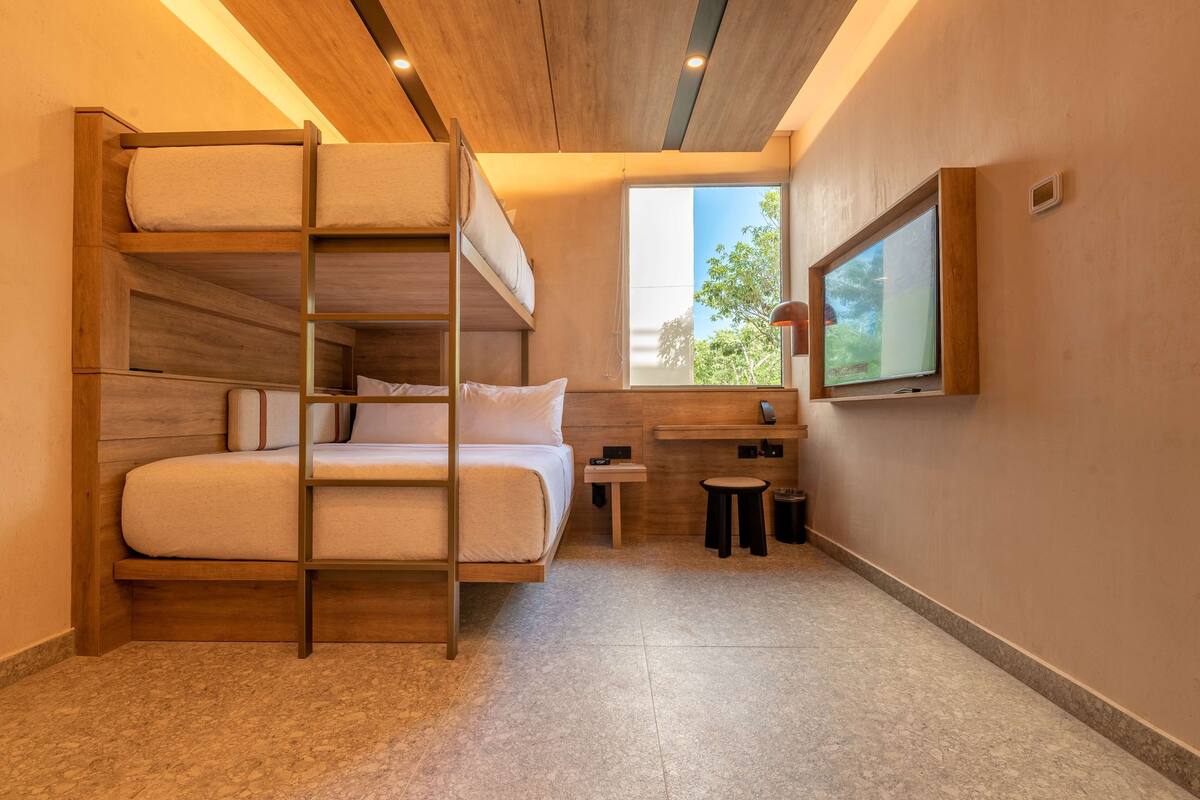 A bunk bed with a wooden frame and white bedding, a small bedside table, a stool, a window with a view of greenery, and a flat-screen TV mounted on the wall.