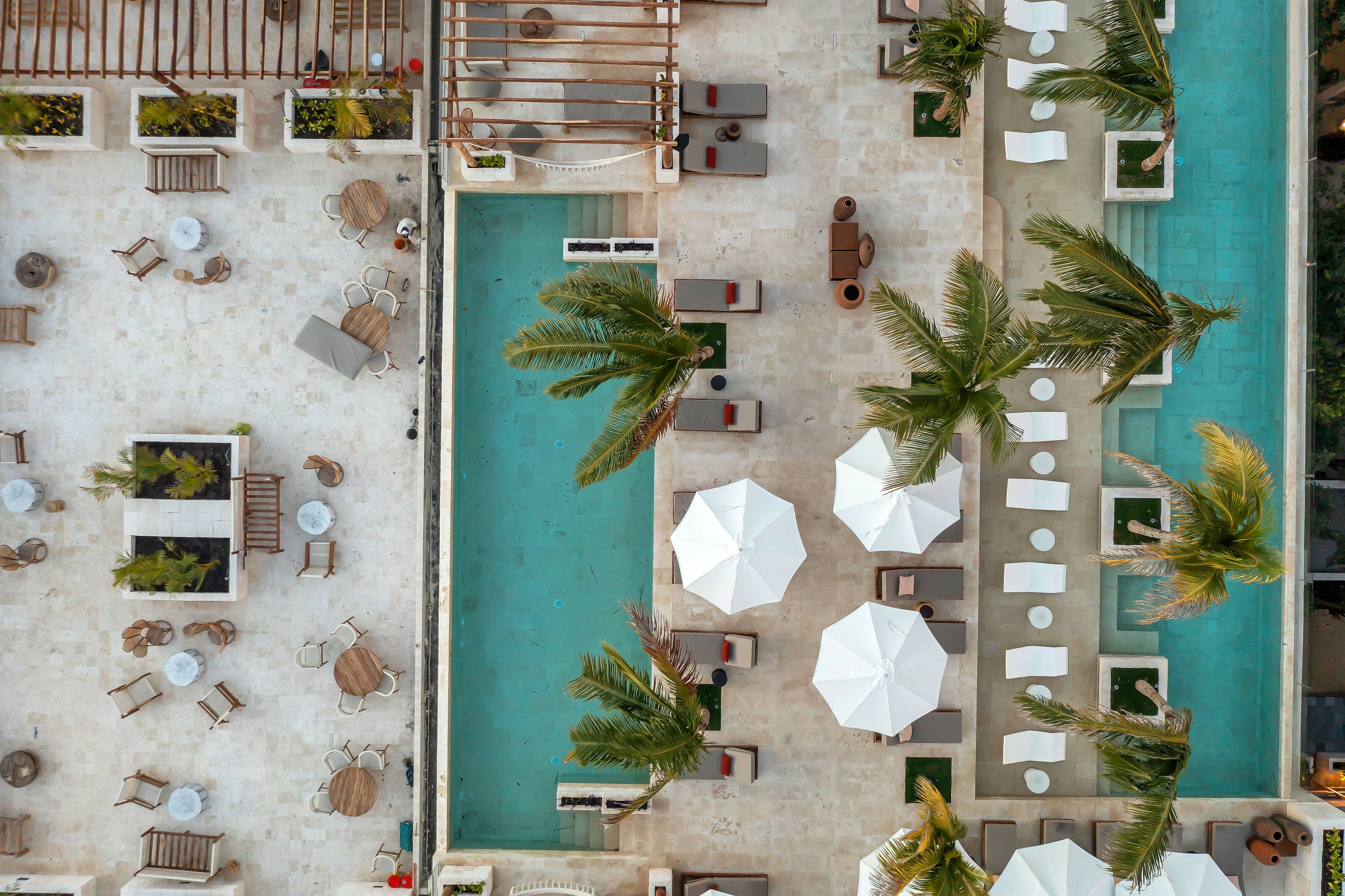 An aerial view of a resort with a swimming pool, lounge chairs, umbrellas, and palm trees.