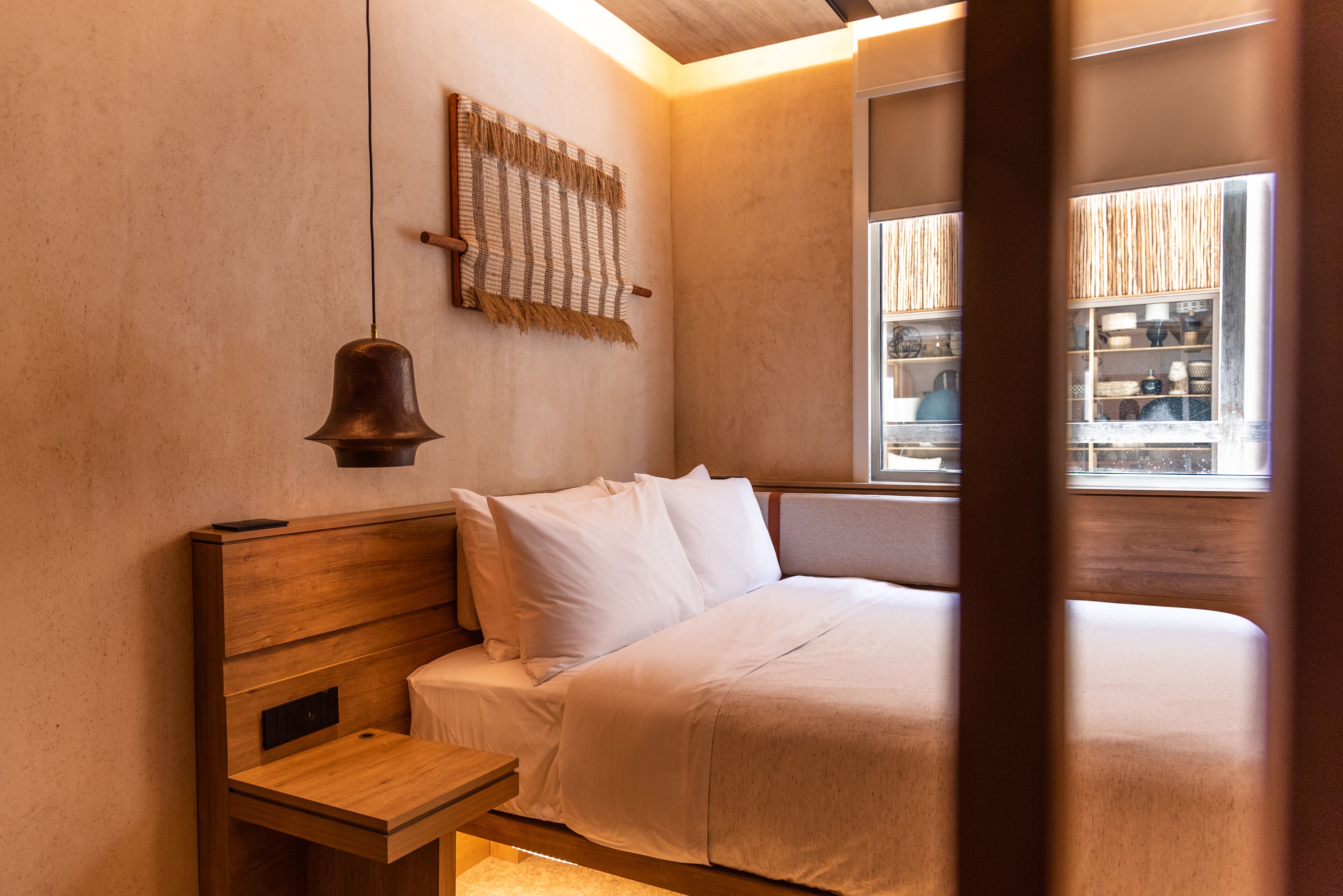 A bedroom with a wooden headboard, a bedside table, a hanging light fixture, and a window with a view of a store.
