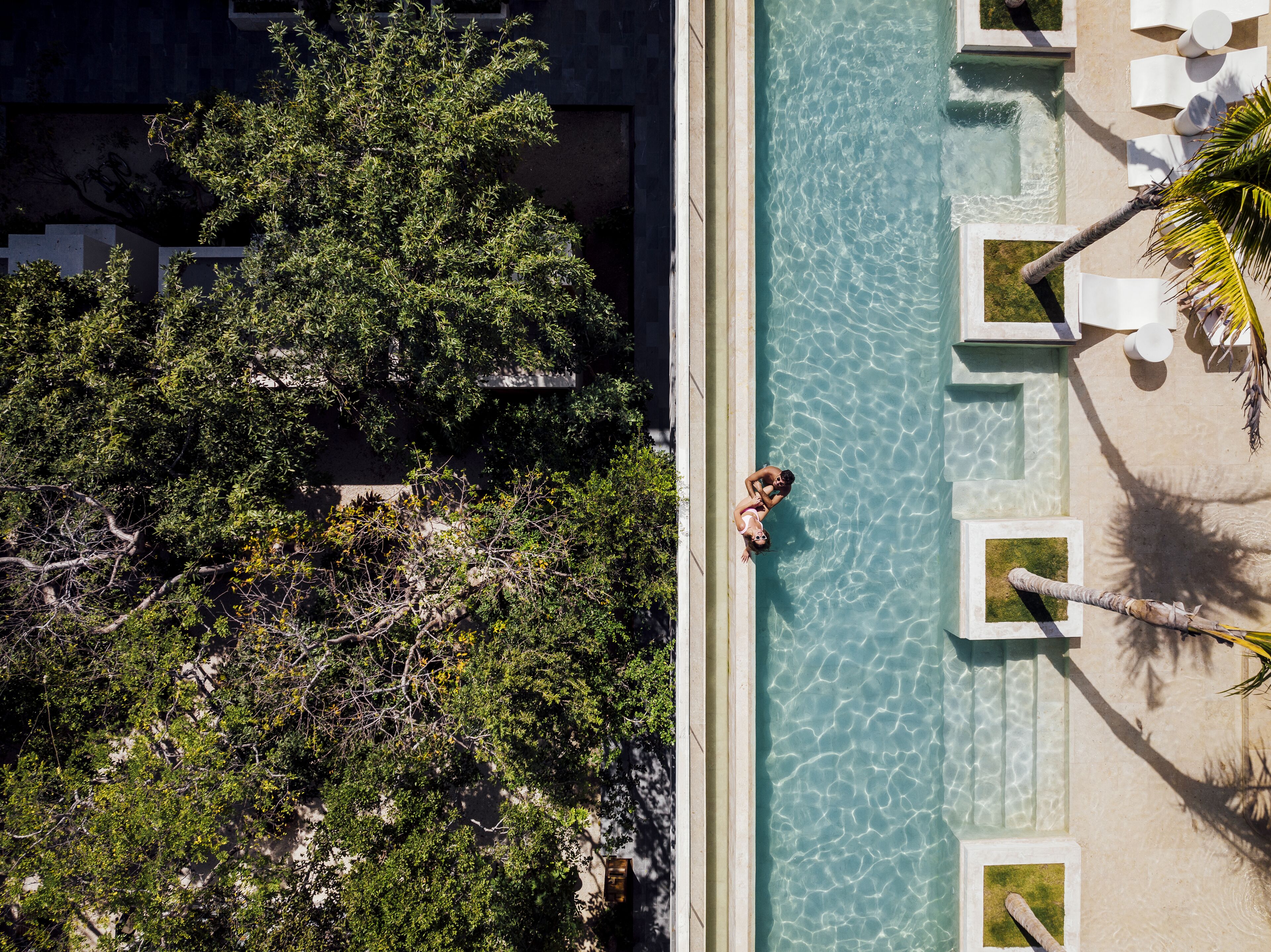 Vue aérienne d’une piscine à l’eau cristalline, entourée d’une végétation luxuriante et de chaises longues blanches.