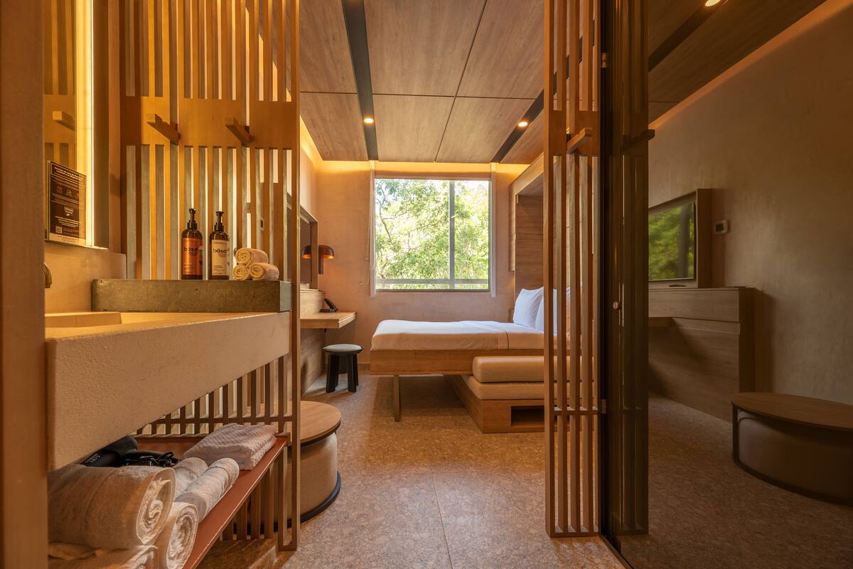 A modern, well-lit room with a wooden ceiling, a large window, a bed, a TV, and a bathroom area with towels and toiletries.