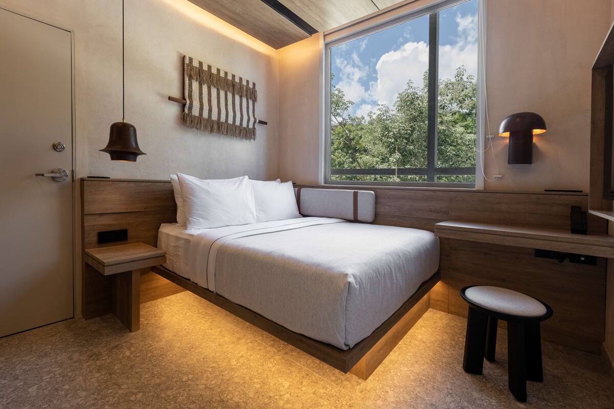 A modern bedroom with a bed, bedside table, and a window with a view of trees.