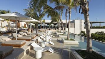 A poolside area with sun loungers and umbrellas, palm trees, and a clear sky.