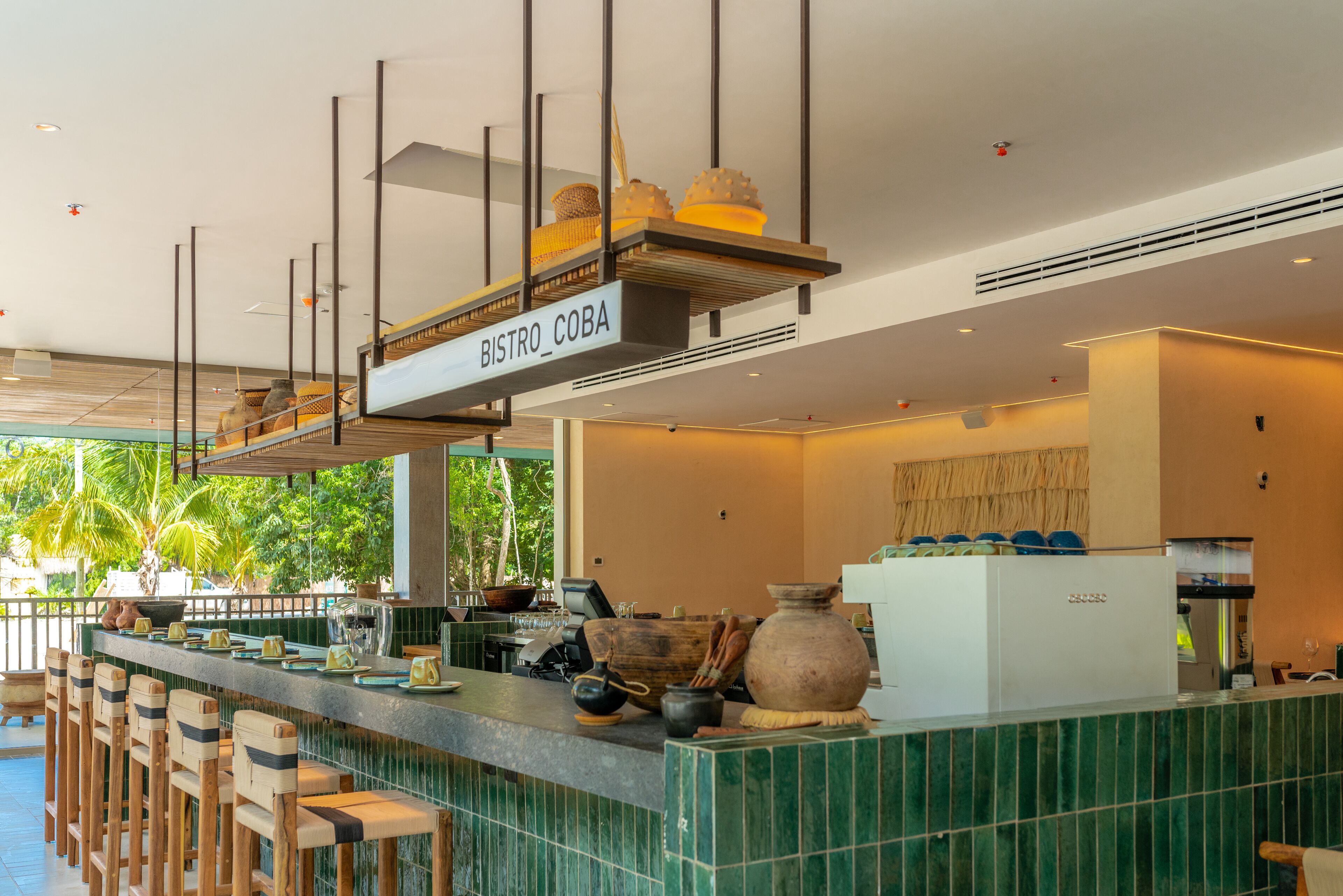 Eine Barbereich mit einem Schild mit dem Schriftzug "BISTRO COBA", hölzernen Barhocker, einer Theke mit Küchenutensilien und Blick auf Grünflächen draußen.