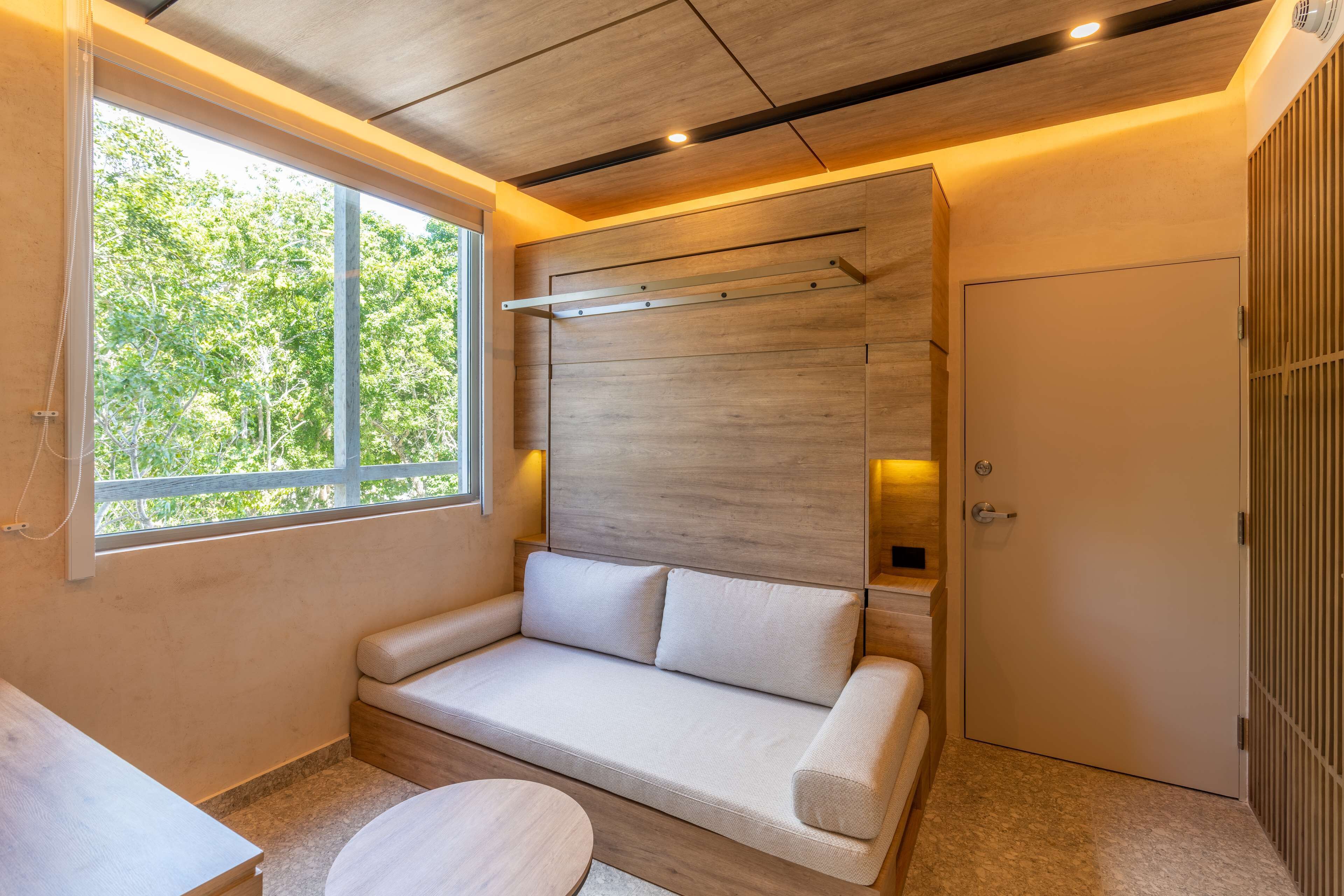 Una habitación moderna con techo de madera, sofá empotrado con cojines, una mesa redonda y ventana con vista a árboles.