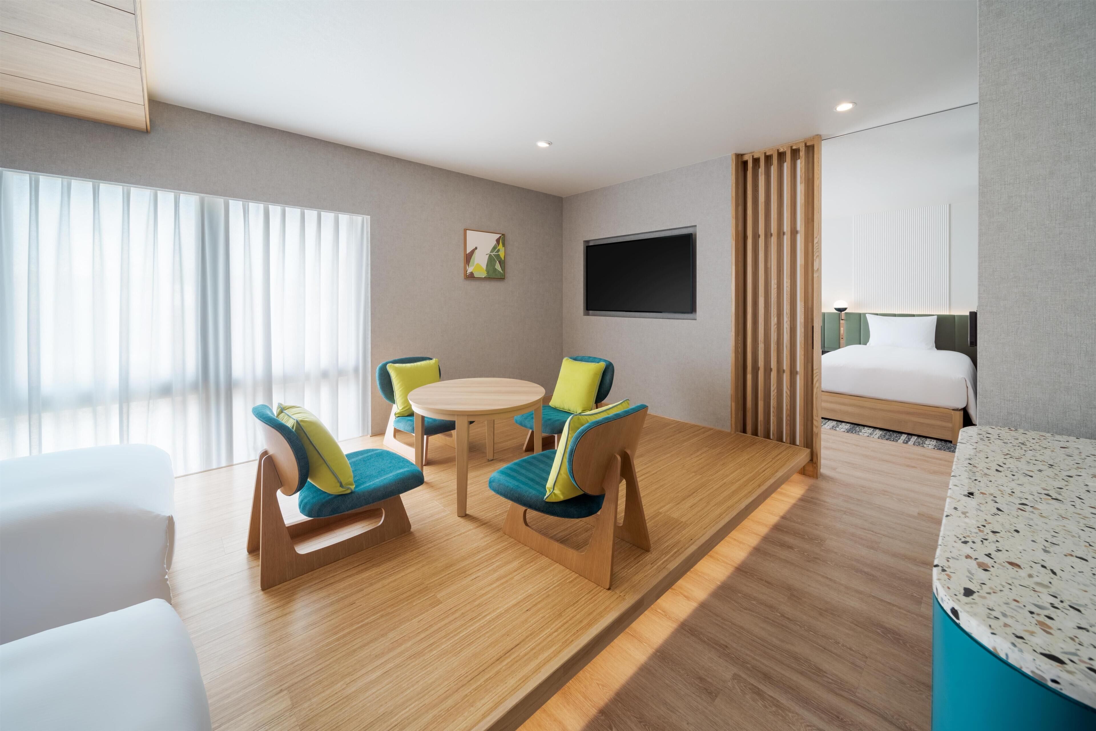 Une chambre d’hôtel moderne avec un plancher en bois, une table ronde avec des chaises, un lit, une télévision et une fenêtre avec des stores.
