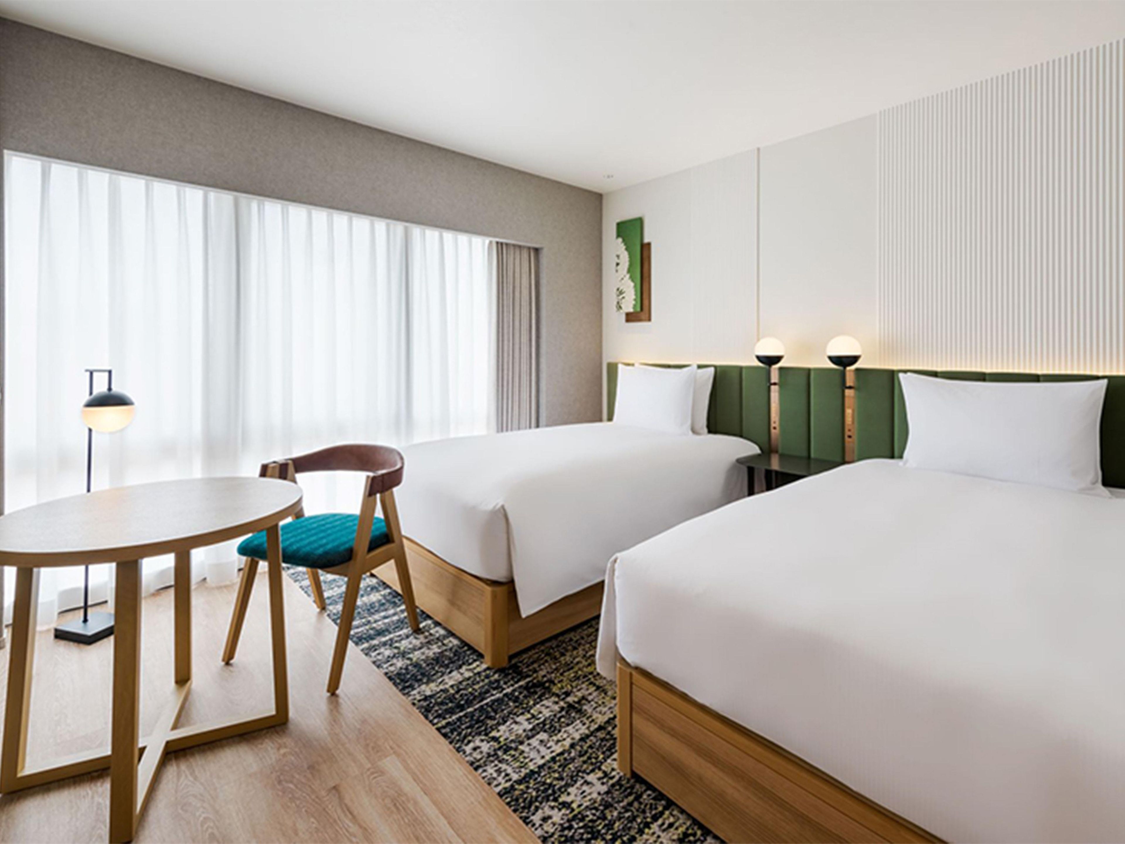 Habitación de hotel con dos camas, una mesita, una silla y un ventanal grande con cortinas.