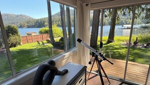 A room with large windows overlooking a lake, a telescope on a wooden deck, and a plush toy on a dresser. - West House! A beautiful lake front home with hot tub and dock. (Shawnigan Lake)