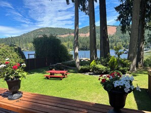 Un patio con una mesa y sillas rojas, rodeado de plantas en macetas y vistas a un lago y colinas boscosas.