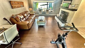 A living room with a leather sofa, a white chair, a coffee table, a fireplace, and a bike in the foreground. - West House! A beautiful lake front home with hot tub and dock. (Shawnigan Lake)