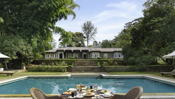 A poolside dining area with a table set for two, surrounded by wicker chairs, overlooking a swimming pool and a house with a stone facade. - The Lavender House by Reveal (Pussellawa)