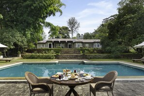 Un espace repas aménagé au bord de la piscine, avec une table pour deux, entouré de chaises en osier, offrant une vue sur la piscine et une maison à la façade en pierre.
