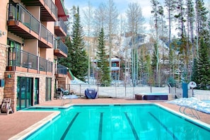 A swimming pool with a view of a mountain resort, a covered patio, and a fenced area with a chair.