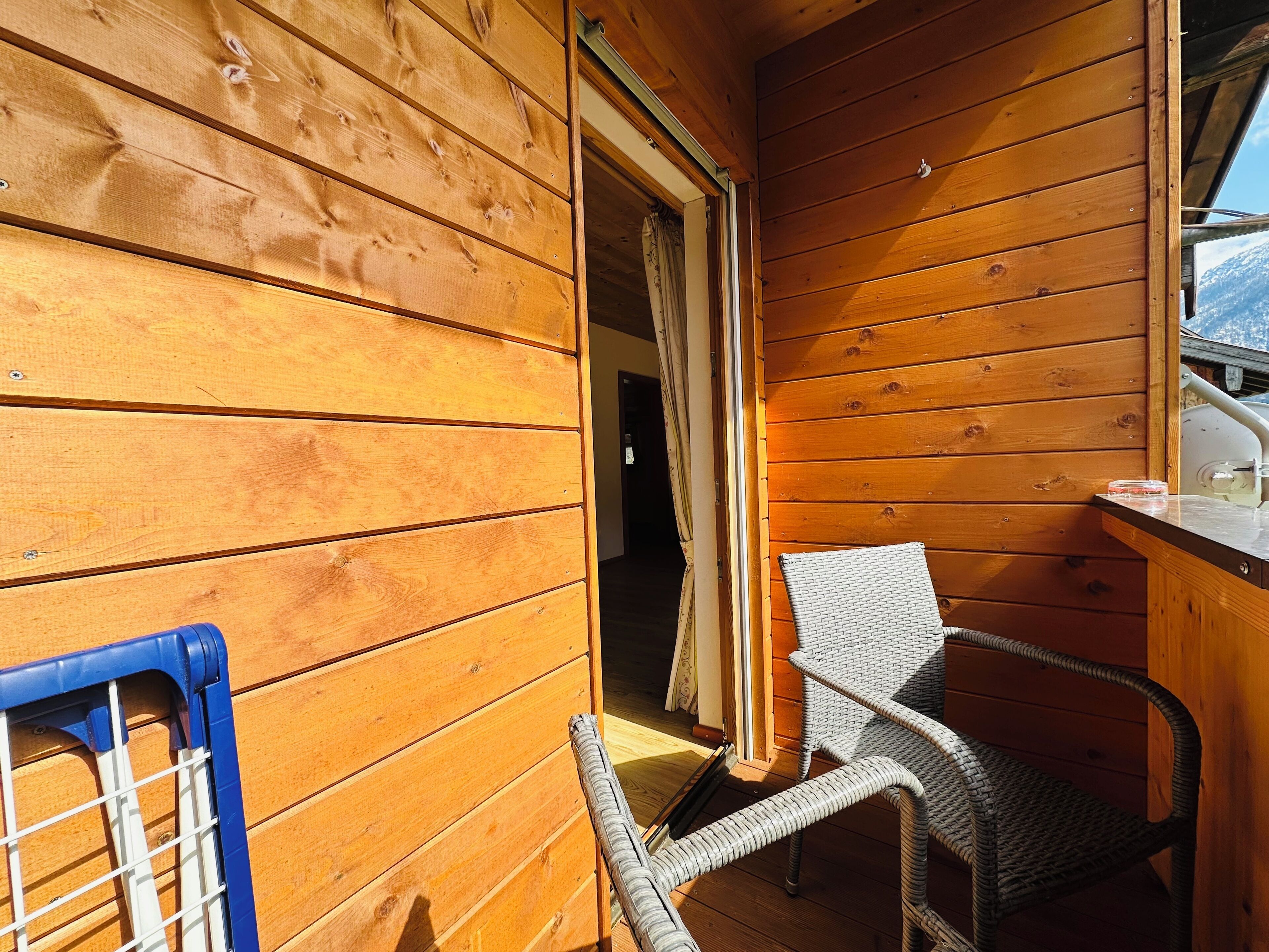 Ein Interieur einer Holzhütte mit einem Korbsessel, einem kleinen Tisch und einem Fenster mit Blick auf schneebedeckte Berge.