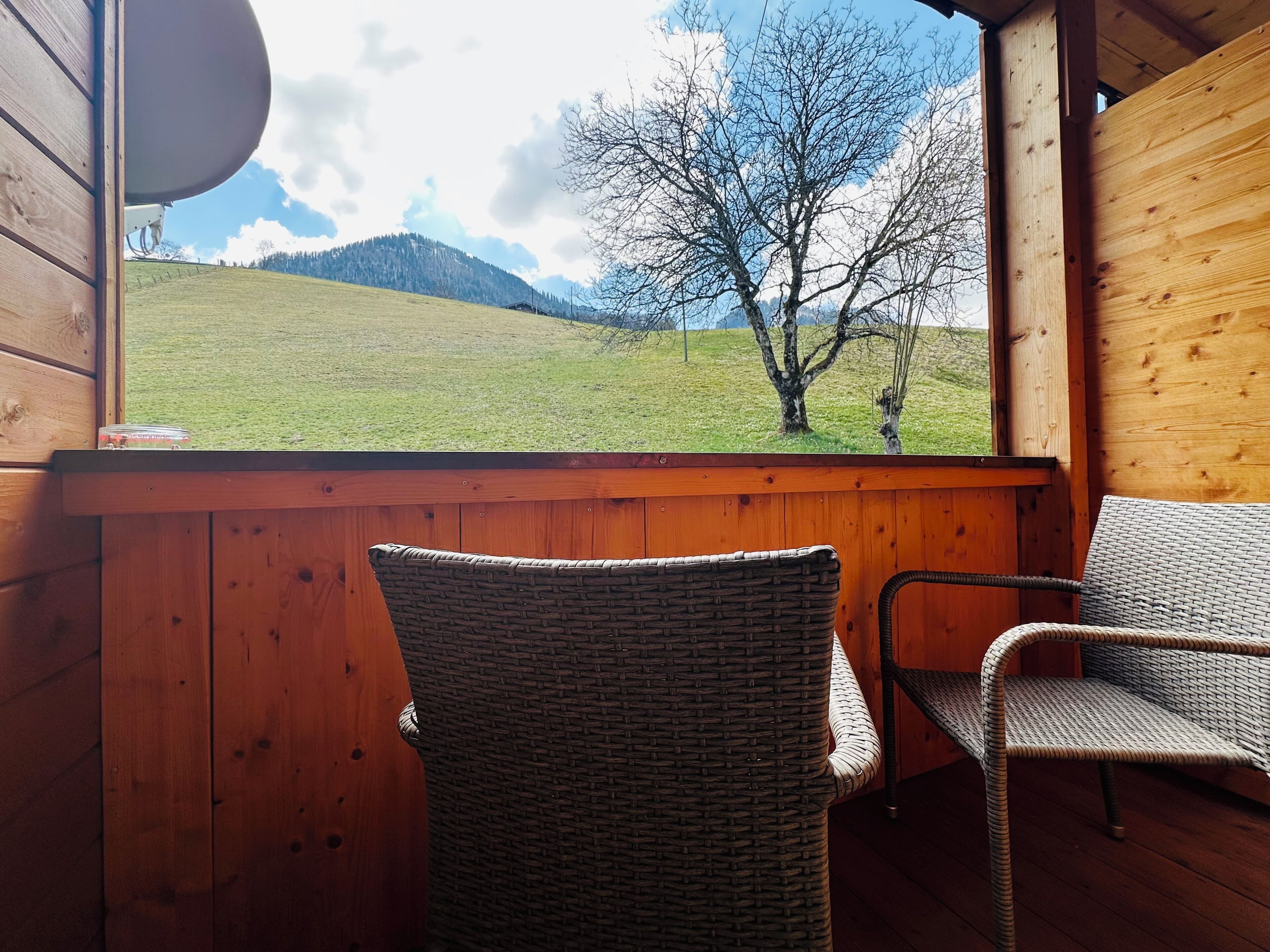 Eine Holzhütte mit Korbsesseln und Blick auf ein Wiesenfeld sowie Berge.