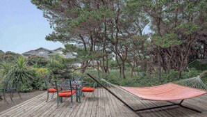 Una terraza de madera con mobiliario exterior, que incluye una mesa y sillas, rodeada de follaje y árboles densos.