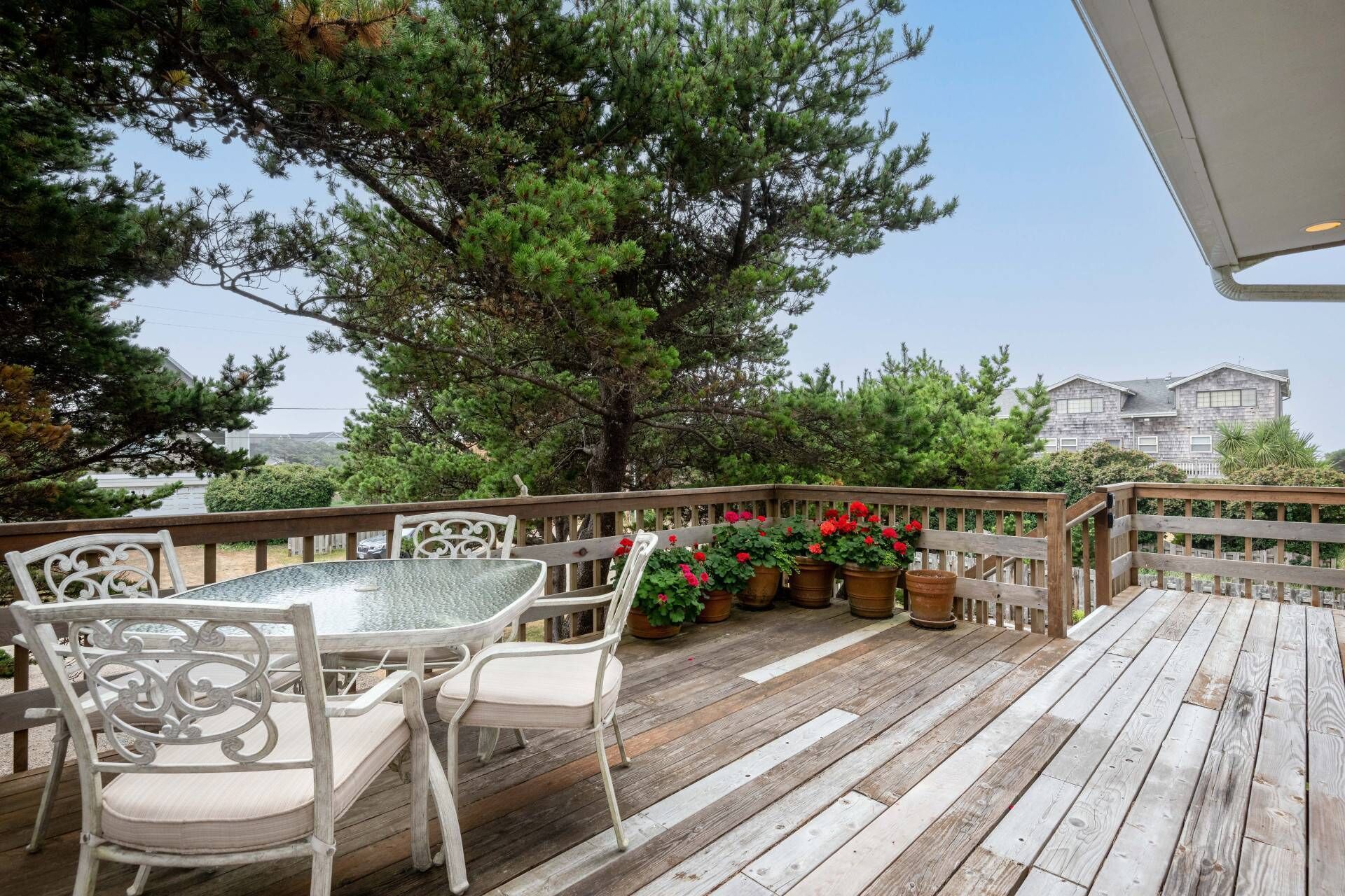 Una terraza de madera con una mesa de vidrio y sillas blancas, rodeada de plantas en macetas y vista a una casa.