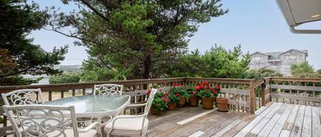 Una terraza de madera con una mesa de vidrio y sillas blancas, rodeada de plantas en macetas y vista a una casa.