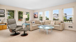 Un salón amplio con un sillón de cuero, un sofá color beige, una mesa de centro de vidrio y ventanas grandes que ofrecen vistas al mar y a los edificios.