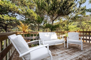 A wooden deck with white outdoor furniture, surrounded by trees. - Spacious Ocean-View Home with Fireplace, Foosball, and Fire-pit, Huge Patio, Steps to the Beach (Cloverdale)