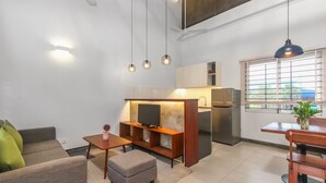 Een moderne woonkamer met een bank, voetenbankje en een eethoek met tafel en stoelen. Er is een keuken met een koelkast en een televisie.