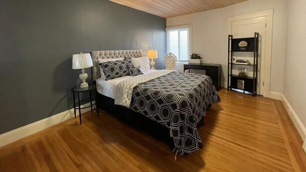 A bedroom with a tufted headboard bed, a bedside table with a lamp, a dark wooden floor, and a built-in shelving unit against the wall.