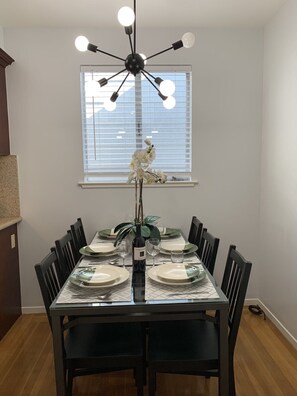 Comedor con mesa de vidrio preparada para seis comensales, rodeada de sillas negras. Un candelabro moderno cuelga arriba, y se ve una ventana con persianas en el fondo.