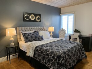 Un dormitorio con cabecera capitonada, una cama con colcha de rayas geométricas en blanco y negro, dos lámparas de noche, una obra de arte en la pared sobre la cama y una ventana con persianas.