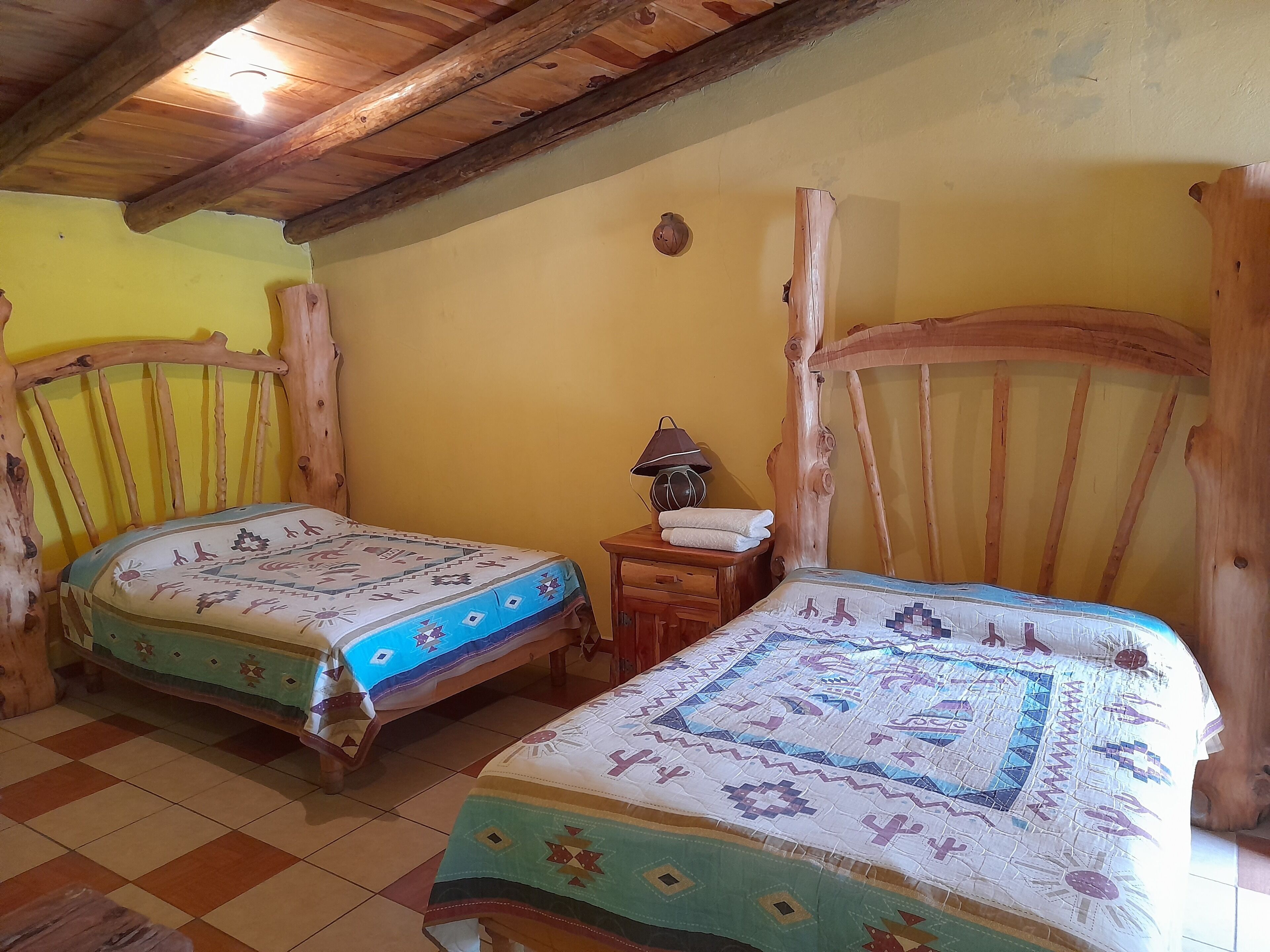 Una habitación rústica con dos camas de madera, una mesita de noche de madera con lámpara y suelo de baldosas.