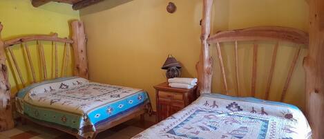 Una habitación rústica con dos camas de madera, una mesita de noche de madera con lámpara y suelo de baldosas.