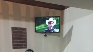 A dining area with a glass table surrounded by chairs, a television mounted on the wall, and a brick accent wall. - Posada Margarito (Urique)