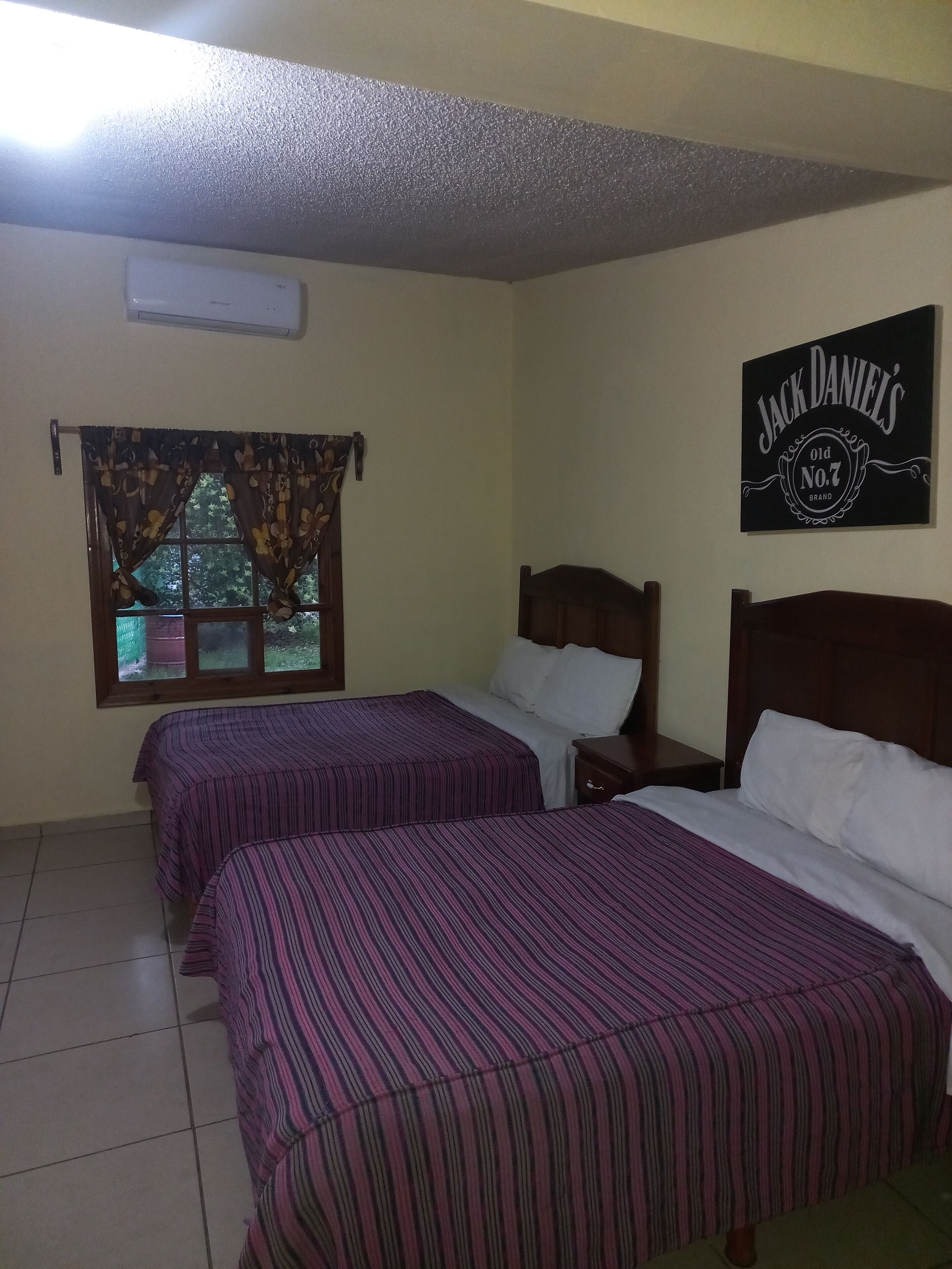 A room with two beds, a window with curtains, a wall-mounted air conditioner, and a sign for Jack Daniel's.