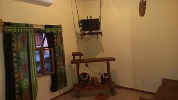 Una habitación con ventana, un banco de madera con objetos decorativos, un equipo de aire acondicionado y un televisor de pared.