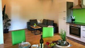 Ein modernes Wohnzimmer mit einer Couch, einem Couchtisch und einem Esstisch, gedeckt mit einer Flasche, Gläsern und Obst. Eine Küche mit Herd und Schränken ist ebenfalls zu sehen.