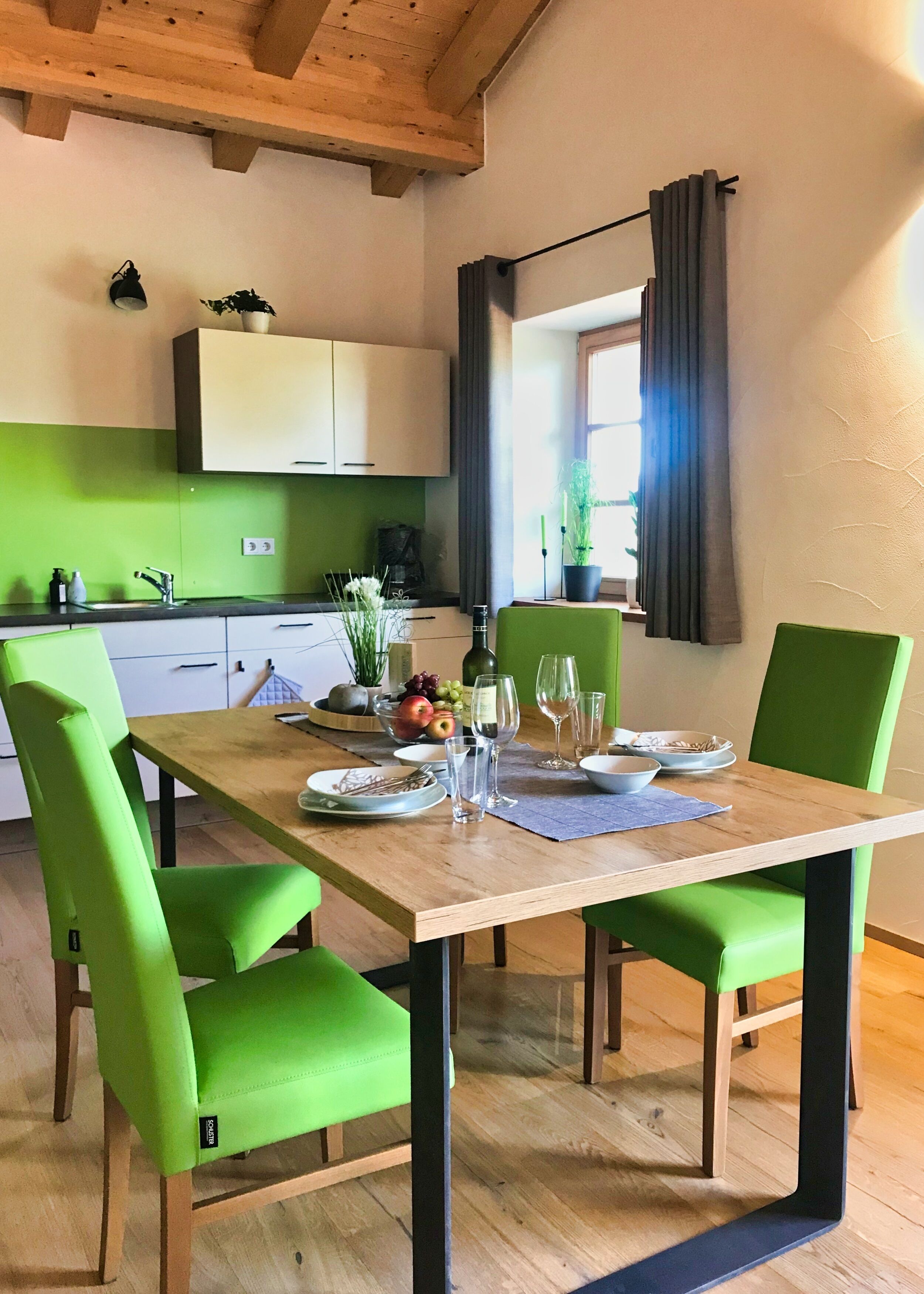 Ein Essbereich mit einem Holztisch, gedeckt für vier Personen, grünen Polsterstühlen und einer Küche im Hintergrund mit grüner Spritzschutzleiste.