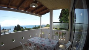 Een overdekt terras met een tafel en stoelen, uitzicht op de zee, en een balkon met een leuning.