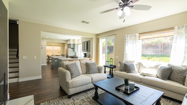 A spacious living room with a sofa set, a coffee table, and a ceiling fan. A kitchen is visible in the background with a dining area.